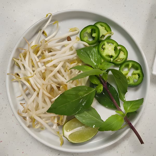 A white plate of fresh pho garnishes: bean sprouts, basil, sliced jalapeños, and a lime wedge.