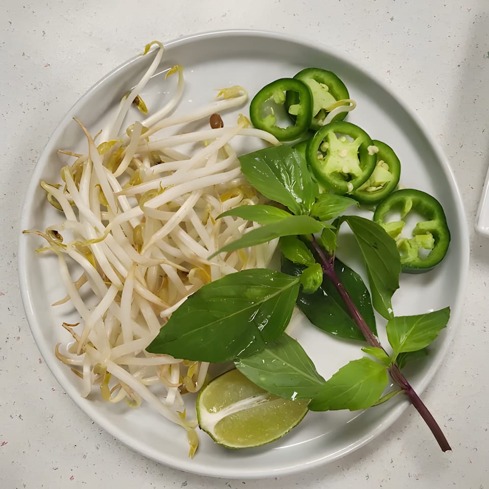 A white plate of fresh pho garnishes: bean sprouts, basil, sliced jalapeños, and a lime wedge.
