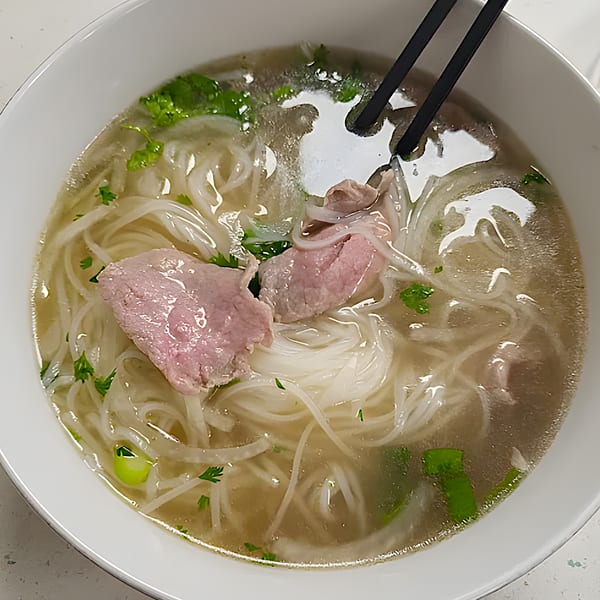 A close-up of Pho W’ Me's Vietnamese pho with beef, noodles, and herbs in a white bowl.