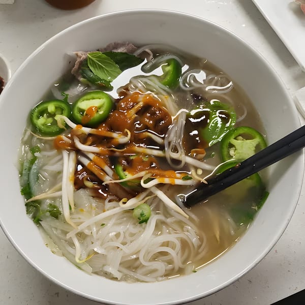 Close-up of Pho W’ Me pho with noodles, meat, jalapeños, bean sprouts, and sauce in a white bowl.